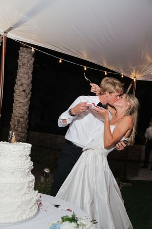 Newlyweds sharing a kiss while cutting their elegant white tiered wedding cake at outdoor reception.