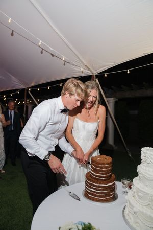 Bride and groom laughing while cutting a rustic naked wedding cake at an outdoor tented reception.