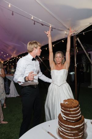 Bride and groom celebrate at wedding reception under tent with string lights, naked cake on table.