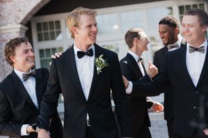 Groom and groomsmen laugh together outdoors in black tuxedos with bow ties and floral boutonnieres.