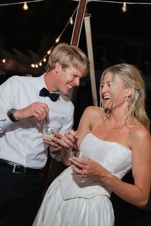 Laughing bride and groom sharing a joyful moment with drinks at their outdoor wedding reception under string lights.