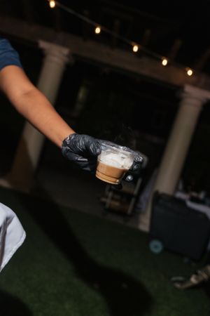 Gloved server holding a steaming hot beverage cup at an outdoor evening wedding reception.