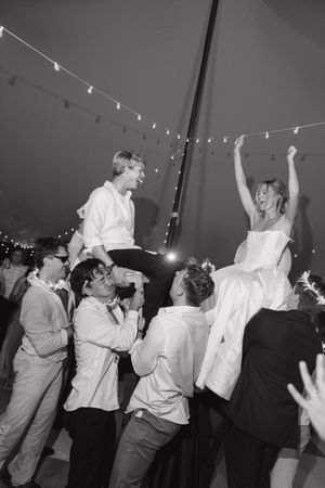 Bride and groom lifted by guests on dance floor at lively outdoor wedding reception under string lights.