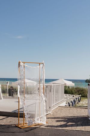 Gold wedding arch draped with white fabric stands on a beachside terrace with ocean views and blue sky.
