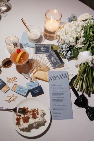 Elegant wedding reception table with blue menu card, cocktail, floral bouquet, candles, and decorative matchboxes.