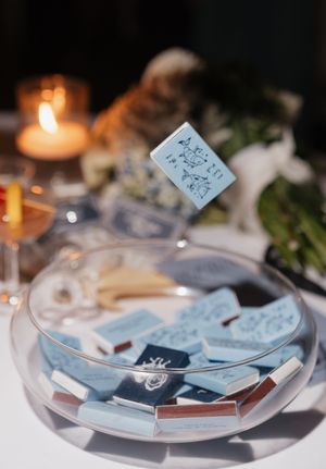 Blue custom matchboxes displayed in a glass bowl as elegant wedding reception favors with candlelight in background.