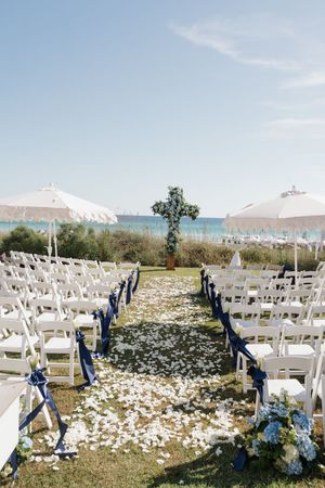 Outdoor beachside wedding ceremony aisle lined with white chairs, navy ribbons, and floral cross altar.