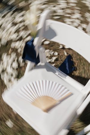 Close-up of white folding chair with wooden hand fan and navy blue ribbon at outdoor wedding ceremony.