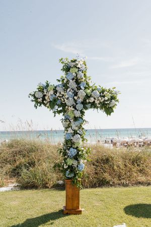 Floral cross decorated with blue hydrangeas and white roses on wooden stand at beach wedding ceremony.