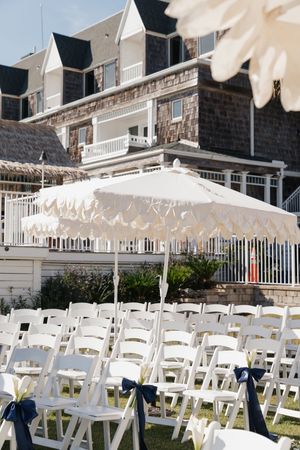 White folding chairs with navy bow decorations arranged on lawn outside a shingle-style coastal venue.
