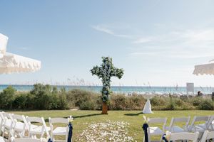 Floral cross wedding altar overlooking the ocean with white chairs and sea oats at beachside ceremony.