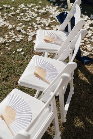 White folding chairs with paper hand fans on seats, set up for an outdoor wedding ceremony on grass.
