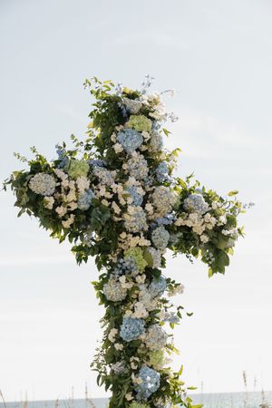 Beautiful floral cross wedding altar decorated with blue hydrangeas and white flowers by the sea.
