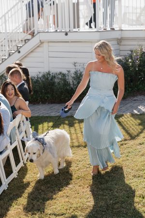 Blonde bridesmaid in light blue strapless dress walks a white dog down the wedding ceremony aisle.