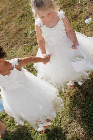 Two flower girls in white dresses holding hands while walking down the grassy wedding aisle.