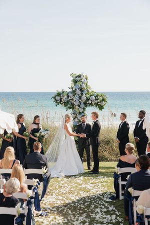 Outdoor beachside wedding ceremony with bride and groom at floral arch, bridesmaids in navy, guests seated on white chairs.