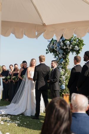 Smiling bride in white gown exchanges vows with groom at outdoor ceremony with blue floral arch.