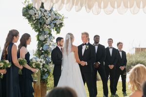 Bride and groom stand at altar with blue floral cross surrounded by bridesmaids and groomsmen outdoors.