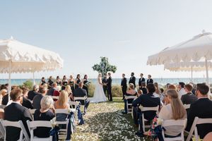 Outdoor beach wedding ceremony with guests seated in white chairs facing couple at a floral altar.