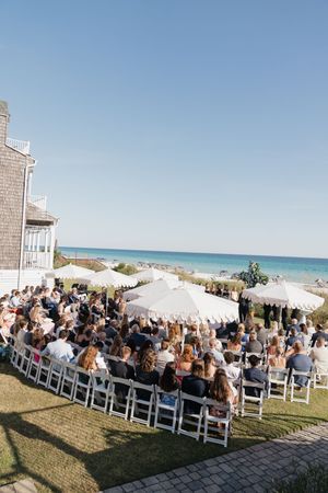 Outdoor beachfront wedding ceremony with guests seated in white chairs under large umbrellas.