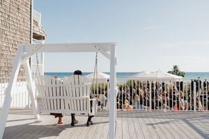 Two wedding guests sit on a white porch swing overlooking a beachside outdoor wedding ceremony on a sunny day.