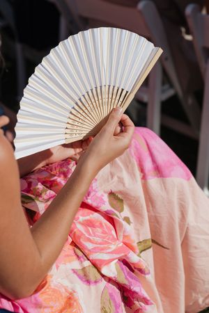 Wedding guest in floral pink dress holding a white folding fan at an outdoor ceremony.