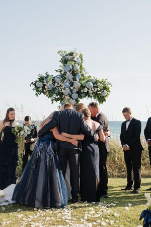 Wedding party shares an emotional group hug at the altar beneath a large floral cross by the ocean.