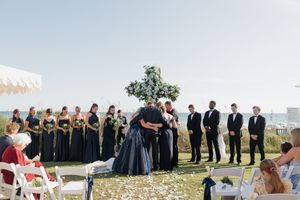 Outdoor wedding ceremony with bridesmaids in navy dresses and groomsmen in black suits, couple embracing at floral arch by the sea.