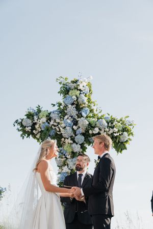 Bride and groom exchange vows before stunning blue and white hydrangea floral cross under clear blue sky.