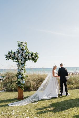 Bride and groom exchanging vows beside floral cross altar at oceanside wedding ceremony.