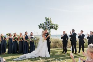 Bride and groom sharing first kiss under floral cross altar with wedding party by the ocean.