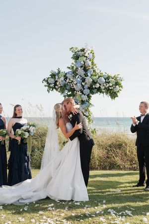 Couple kissing at altar beneath blue and white floral cross with bridesmaids in navy dresses.