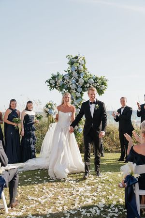 Joyful bride and groom walk back down aisle after ceremony beneath floral cross arch by the ocean.
