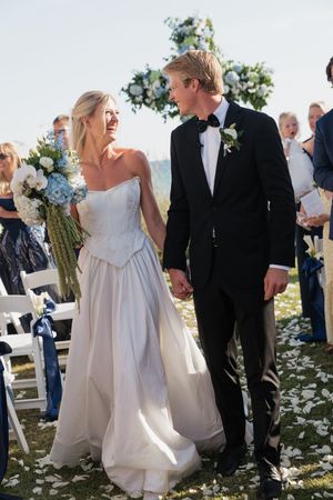 Radiant newlyweds walk down the aisle hand-in-hand past seated guests with a floral arch and blue hydrangea bouquet.