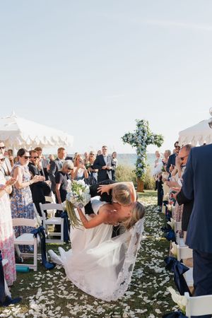Newlyweds share a dip kiss walking down the aisle as guests toss petals at outdoor ceremony.