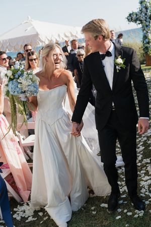 Laughing bride and groom walk hand-in-hand down the aisle after outdoor seaside wedding ceremony.