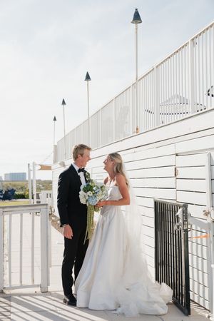 Bride and groom sharing a smile on a white waterfront boardwalk holding a blue floral bouquet.