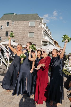 Bridesmaids in navy gowns celebrating with raised bouquets outside a coastal wedding venue on a sunny day.