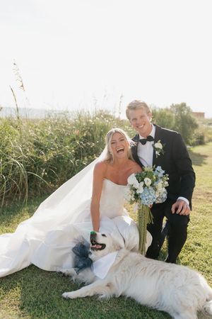 Laughing bride and groom in wedding attire pose outdoors with their large white dog near coastal sea grass.