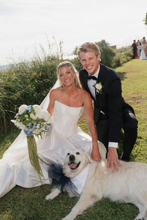 Smiling bride and groom pose with their white golden retriever dog at outdoor wedding venue.