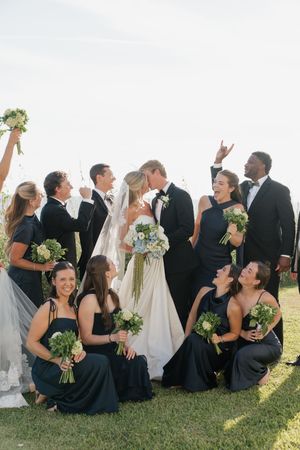 Wedding party celebrating as bride and groom kiss on a sunny lawn, bridesmaids in navy dresses holding green bouquets.