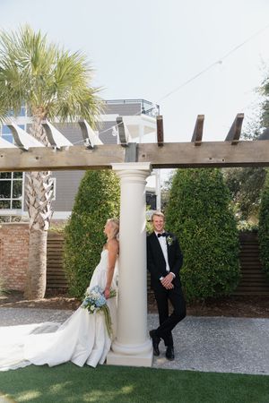Bride and groom pose by a white column under a wooden pergola surrounded by lush greenery at their wedding.