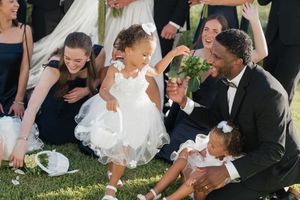 Wedding party member in tuxedo plays with two young flower girls in white dresses on green lawn.