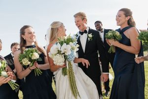 Bride and groom walk with wedding party in navy attire holding white and blue bouquets outdoors.