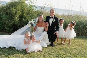 A bride and groom pose with three young flower girls on a grassy hillside during an outdoor wedding ceremony.