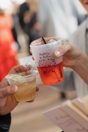 Two hands clinking personalized wedding cups with colorful drinks at a celebration event.