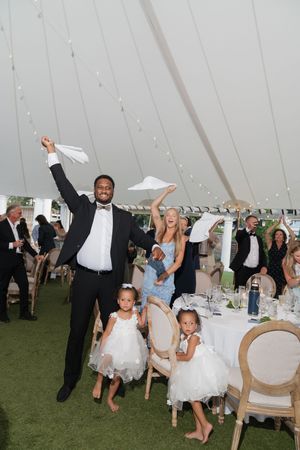 Joyful wedding reception under a white tent with guests celebrating, flower girls in white dresses, and festive string lights.
