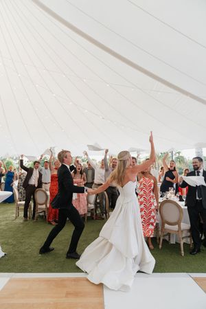 Bride and groom celebrate first dance under white tent with cheering wedding guests at outdoor reception.