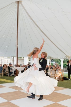 Groom spinning bride in her flowing white gown on checkered dance floor at wedding reception.