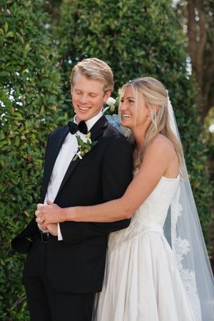 Smiling bride and groom embracing outdoors in front of lush green hedges after ceremony.